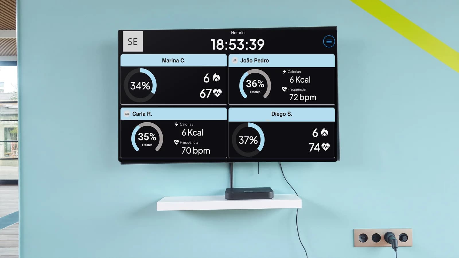 TV na parede da academia exibindo o Vektor Pulse, com o receptor na prateleira abaixo